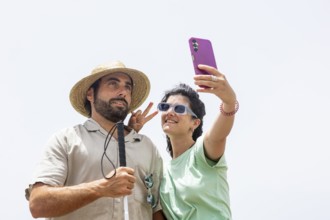 A blind man and a woman smile while taking a playful selfie by the sea. The woman makes a peace