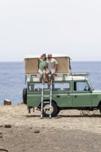 A blind man and a woman sit on the rooftop tent of a motorhome, enjoying a coastal view while