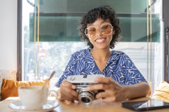 A cheerful woman at a cafe, holding a vintage camera, with a cup of coffee in the foreground.