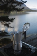 A moka pot sits on a camping stove beside a tranquil Norwegian lake, surrounded by lush mountains