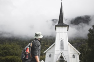 A traveler with a backpack and hat stops in front of a picturesque white Norwegian church,