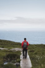A traveler wearing a red jacket and backpack walks along a wooden path overlooking the Norwegian
