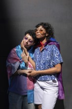 A multiethnic LGBTQ+ couple stands closely together, wrapped in colorful flags. Both wear