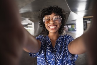 Happy woman with curly hair and sunglasses takes a selfie in an industrial setting. She wears a