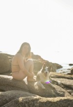 A woman in a bikini sits beside her happy dog on sunlit rocks by the sea The peaceful setting