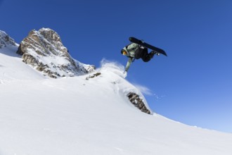 A snowboarder executes an aerial trick on a snowy slope under a vibrant blue sky. The dynamic