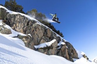 A snowboarder catches air while leaping off a rocky cliff, against a vibrant blue sky. The scene