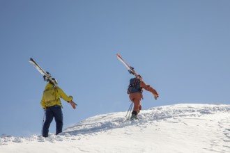 Two skiers in colorful gear hike up a snowy slope, carrying their skis The clear blue sky contrasts