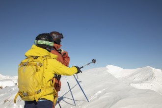Two freeride skiers stand atop a snow-covered peak, gazing into the vast mountain range under a