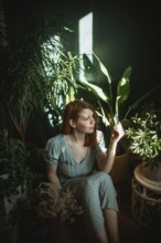 A woman in a green dress sits surrounded by lush indoor plants, holding a small card. Sunlight
