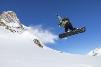 A snowboarder performs a jump against a clear blue sky, with snow spraying behind. The stunning