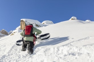 A snowboarder, wearing a red backpack, climbs a snowy mountain with a snowboard. The scene captures