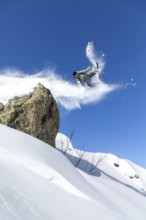 A snowboarder performs an aerial trick off a snowy cliff against a clear blue sky, capturing the