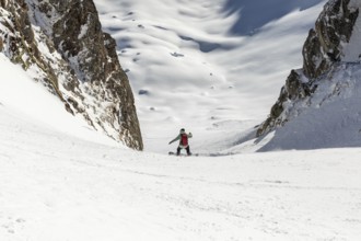 A snowboarder carves down a steep, snow covered slope between rocky peaks. The bright snow