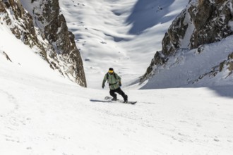 A snowboarder skillfully descends a steep, snowy mountain slope surrounded by rugged cliffs. The