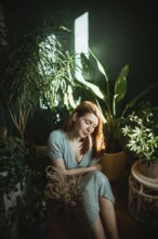 A woman in a light dress calmly sits surrounded by lush indoor plants. Sunlight filters through,