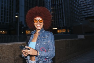 Fashionable African descendent woman with vibrant afro hair smiles while checking her smartphone in