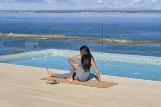 A woman in athletic wear performs yoga on a mat by an infinity pool with a stunning ocean backdrop,