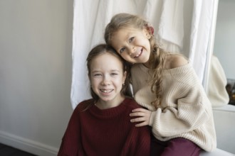 Two sisters wearing cozy sweaters smile warmly at the camera The younger sibling leans