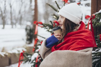 Young woman wrapped in a red scarf, holding a coffee cup, and smiling, seated near a festive
