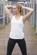 A young blond woman wearing a white tank top and black leggings is stretching her arms above her