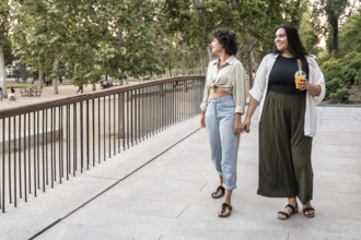 A lesbian couple walks hand in hand through a park, enjoying a leisurely stroll. One holds a drink,