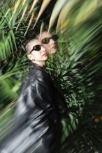 A stylish lesbian woman wearing sunglasses and a leather jacket stands amidst lush green foliage.