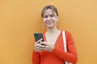 A woman stands against an orange wall in Warsaw, intently using her smartphone Dressed in a red top