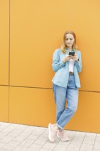 A woman leans against a bright orange wall in Warsaw, focused on her phone She wears a blue shirt,