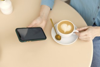 A woman relaxes in a Warsaw cafe, holding a smartphone and a cup of coffee with latte art The cozy
