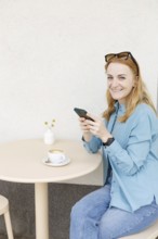 A smiling woman enjoys coffee while using her smartphone at a cafe in Warsaw She wears casual