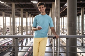 A young traveler in a vibrant blue shirt leans casually against a railing at a train station,