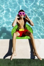 Young teenage boy sits on a green float in a pool, wearing vibrant red shorts, flower-shaped pink
