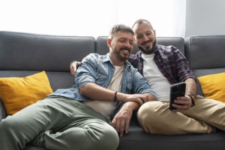 A joyful gay couple sits comfortably on the sofa, smiling as they look at a smartphone together in