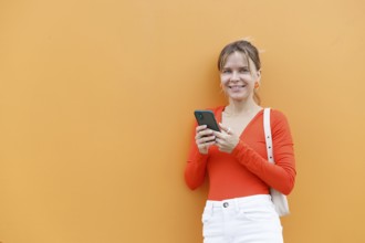 A woman wearing a red top stands against an orange wall, holding a smartphone The vibrant colors