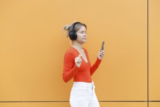 A young woman wearing headphones, joyfully listening to music and holding a smartphone She stands