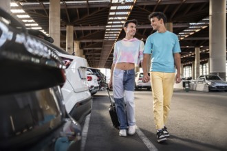 A gay couple walks hand in hand through a sunny parking area filled with cars. Both are smiling and