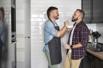 A joyful gay couple shares a moment in the kitchen, one feeding the other with a fork while smiling
