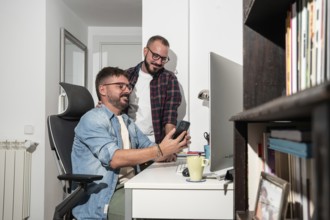 A joyful gay couple shares a moment at their home office. One man sits at a desk, holding a phone,