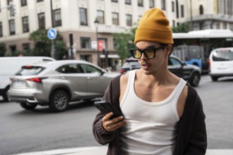 A Colombian man stands on a busy city street, wearing stylish sunglasses and a beanie as he checks