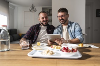 A gay couple enjoys breakfast together at a table, sharing a tablet and smiling. Fresh fruit,