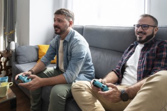 A gay couple is sitting on a sofa, joyfully playing video games together in a bright and cozy