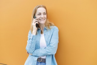 Smiling woman stands against a vibrant orange wall in Warsaw, chatting on a smartphone She wears