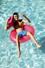 Young teenage boy enjoys a sunny day in a swimming pool, lounging on a pink inflatable ring. He
