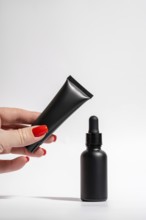 Cropped unrecognizable female hand with red manicure holding a matte black tube and dropper bottle