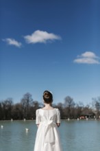 A young girl in a white First Communion dress stands by a tranquil lake, gazing at the serene view.