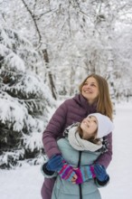 A joyful mother embraces her daughter during a snowy winter day. They both wear colorful winter