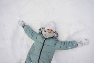 A child joyfully lies in the snow, wearing a green coat, white hat, and scarf. The scene captures