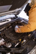 A person in winter clothing checks car fluids in snowy Swedish Lapland. Hood open, engine exposed,