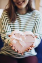 Cropped unrecognizable woman in a striped top holding a heart-shaped gift box adorned with a white